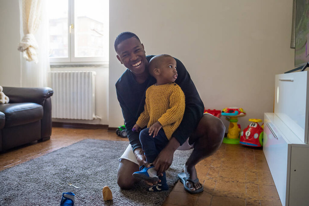 Vader die thuis op de grond zit en schoenen aantrekt aan zijn peuter - alleenstaande ouder die zich omkleedt aan zijn baby - dagelijkse routine, band, familieconcept - Foto, afbeelding