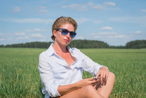 beautiful woman in a white shirt sits in a summer field on the grass with a phone in her hands - Fotoğraf, Görsel