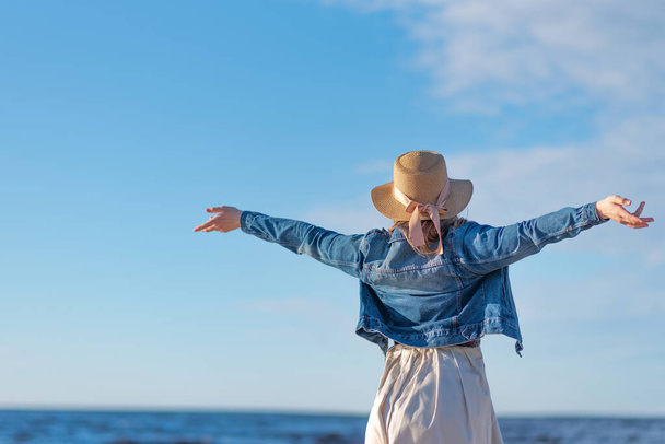 woman wearing a beige wide dress, denim jacket and straw hat stands on the beach with arms wide open - Foto, imagen