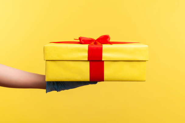 Profile side view closeup of human hand in blue surgical gloves holding yellow gift box. indoor, studio shot, isolated on yellow background. - Photo, image