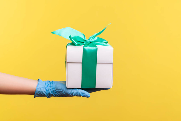 Profile side view closeup of human hand in blue surgical gloves holding white gift box. sharing, giving or delivery concept. indoor, studio shot, isolated on yellow background. - Foto, immagini