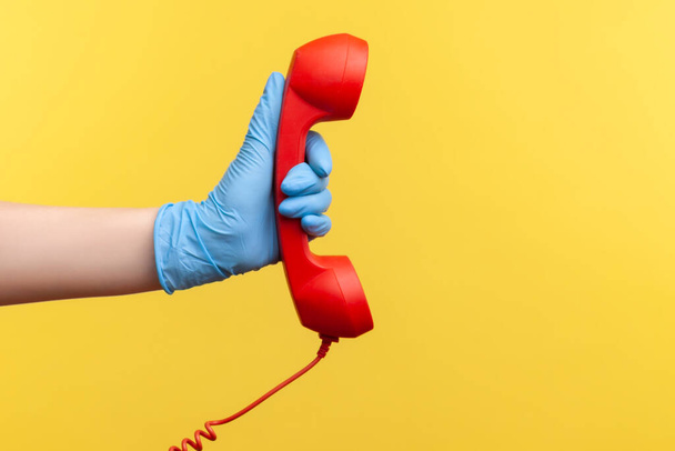 Profile side view closeup of human hand in blue surgical gloves holding and showing red call telephone handset receiver. indoor, studio shot, isolated on yellow background. - Fotoğraf, Görsel