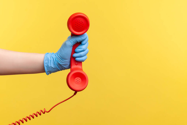 Profile side view closeup of human hand in blue surgical gloves holding and showing red call telephone handset receiver. indoor, studio shot, isolated on yellow background. - Фото, изображение