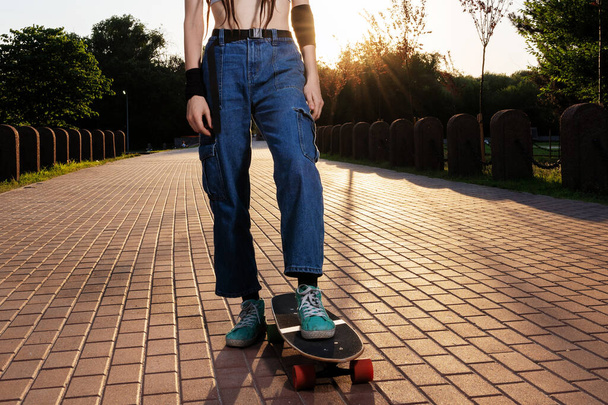 slank meisje met dreadlocks in jeans en een top berijdt een longboard bij zonsondergang. mooie tiener meisje skater. zomervakantie - Foto, afbeelding