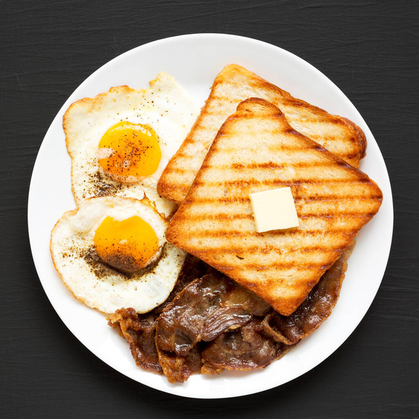 Hausgemachtes gesundes Sunnyside Eggs Frühstück auf einem weißen Teller auf schwarzer Oberfläche, Draufsicht. Flach liegend, über Kopf, von oben. Nahaufnahme. - Foto, Bild