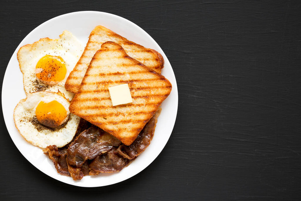 Hausgemachtes gesundes Sunnyside Eggs Frühstück auf einem weißen Teller auf schwarzer Oberfläche, Draufsicht. Flach liegend, über Kopf, von oben. Raum für Text. - Foto, Bild