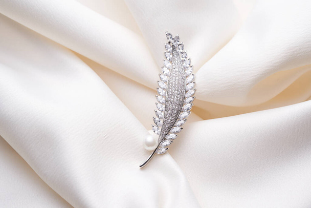Silver brooch in the shape of a feather, with small diamonds and white pearl isolated on a delicate background of feathers.  - Photo, Image
