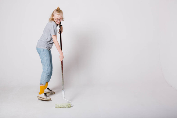 Girl painter paint the floor white with a construction roller. Blonde painter performs painting works. - Photo, Image