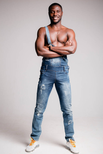 Dark-skinned African-American man with naked torso in denim pants with suspenders crossed his arms on his chest. - Фото, зображення
