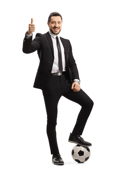 Full length portrait of a man in a black suit with his leg on a soccer ball showing thumbs up isolated on white background - Foto, Imagen