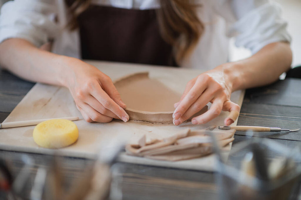 Ремесленные руки соединяют глиняные детали из сырой глины, мастерские пальцы работают с кусочками глины, женщины сидят в мастерской за столом - Фото, изображение