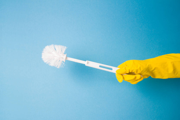 toilet brush in hand on a blue background, a hand in a yellow rubber glove holding a white plastic toilet brush - Фото, изображение
