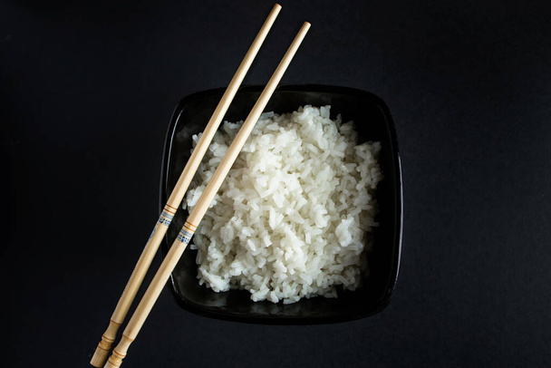 Bowl with boiled rice on a black background. Asian food and bamboo chopsticks. - Φωτογραφία, εικόνα