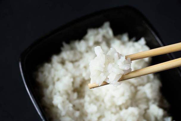 Schüssel mit gekochtem weißen Reis auf schwarzem Hintergrund. Asiatisches Essen und Bambusstäbchen. - Foto, Bild