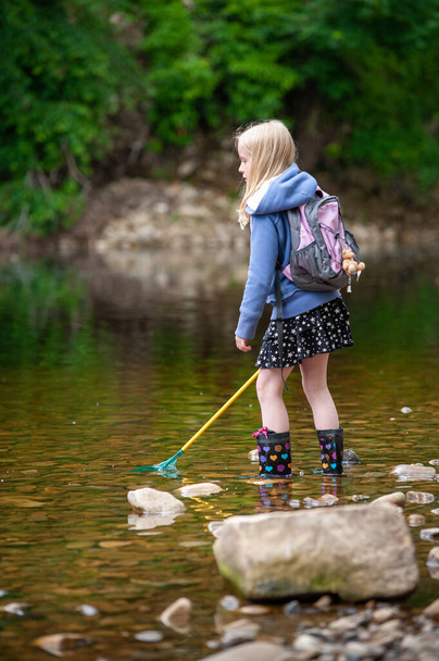 Nuori blondi tyttö reppu ja kalaverkko kahlaa matalassa vedessä joen yllään wellies - Valokuva, kuva