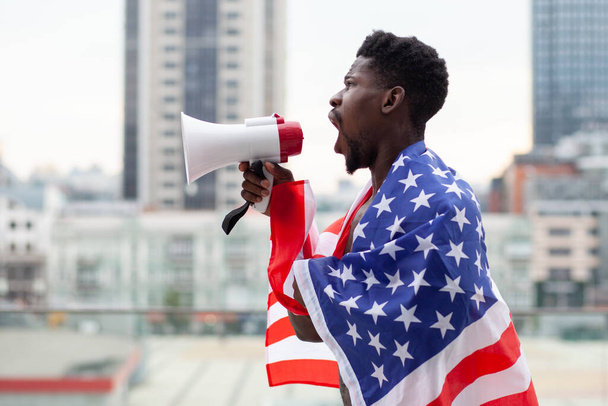 L'uomo afroamericano con la bandiera degli Stati Uniti grida in un megafono e protesta sullo sfondo della città, le vite nere contano il concetto - Foto, immagini