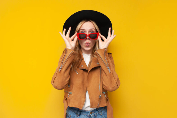 young shocked girl in autumn clothes and glasses is surprised at a yellow isolated background, a woman takes off her glasses and says wow - Photo, Image