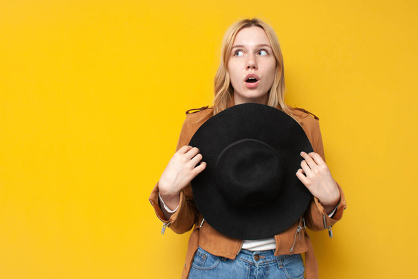 νεαρή έκπληκτη κοπέλα με φθινοπωρινά ρούχα κρατά ένα καπέλο και κοιτάζει copyspace σε ένα κίτρινο απομονωμένο φόντο - Φωτογραφία, εικόνα