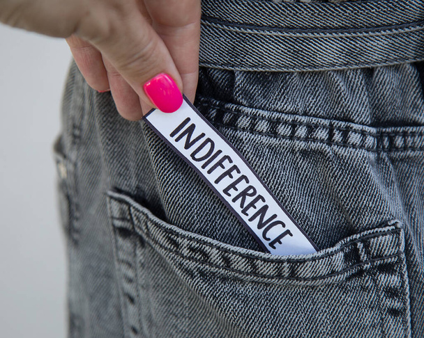 Красивая женская рука с ярким маникюром берет из кармана джинсов бумагу с надписью "Безразличие". Фото для статьи по психологии, коучинг для сайта, социальные сети
 - Фото, изображение