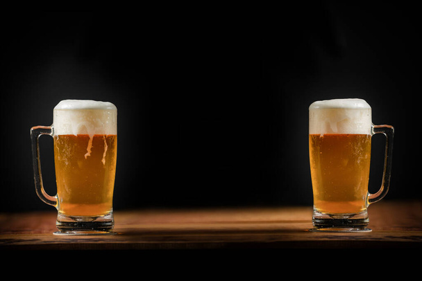 two cold mugs with beer, with overflowing foam, on wooden table and dark background, space for writing - Photo, Image