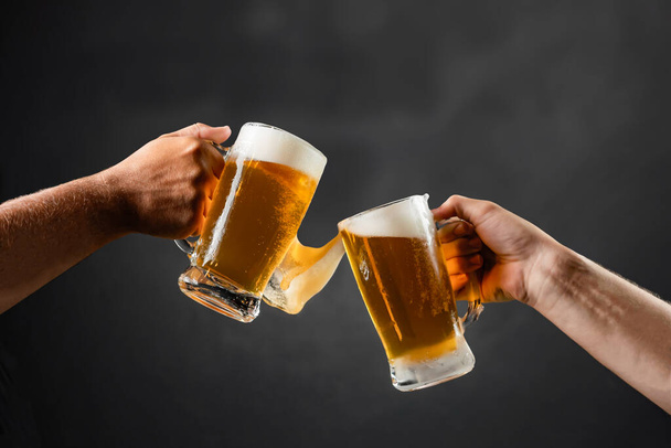 two hands with beer mugs, toasting in celebration, with overflowing foam, dark background and space for writing - Photo, Image