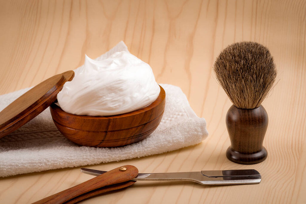 Male cosmetic products and supplies used by men to shave concept with a straight razor, towel, shaving brush and foam on wood background - Photo, Image