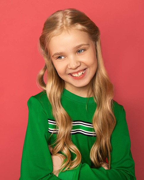 Photo of beautiful blonde girl of 8-10 years old with blue eyes, hair arranged in curls in bright clothes on a pink background shyly smiles and laughs. Studio Close up portrait. Positive emotions - Fotoğraf, Görsel