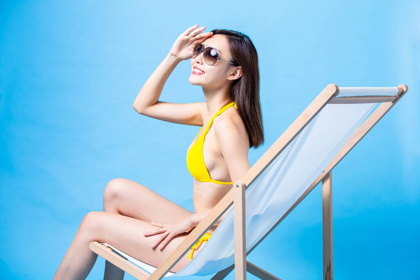 summer sunblock and travel concept - asian bikini girl lying on beach chair with her hand block sunshine isolated blue background - Photo, image