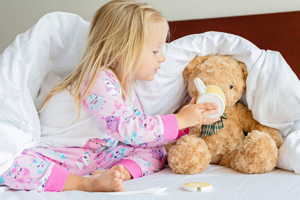 Cute little girl with blonde hair sitting on the bed with stuffed teddy bear. Happy childhood. Stay at home during coronavirus covid-19 pandemic quarantine concept. - Fotografie, Obrázek