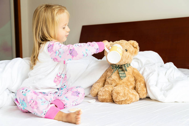 Cute little girl with blonde hair sitting on the bed with stuffed teddy bear. Happy childhood. Stay at home during coronavirus covid-19 pandemic quarantine concept. - 写真・画像