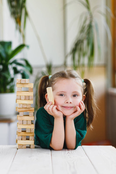 Portret pięknej uśmiechniętej dziewczynki w zielonej sukience z kucykami na głowie trzymającej drewniany blok i drewnianą wieżę stojącą na stole. - Zdjęcie, obraz