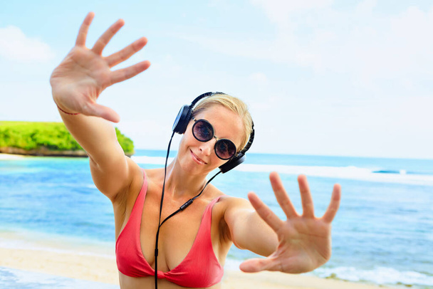 Young positive girl in headphones and sunglasses listening music and dancing with fun at tropical beach party. Travel family lifestyle, recreational activities at summer cruise vacations. - Fotoğraf, Görsel