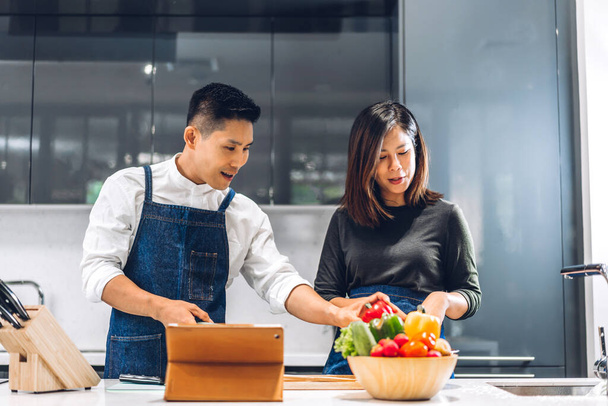 Genç Asyalı çift masada taze sebze salatasıyla yemek yaparken eğleniyor. Mutfakta lezzetli bir yemek hazırlamak için tablet bilgisayarlı ve internette yemek tarifi arayan mutlu çift. - Fotoğraf, Görsel