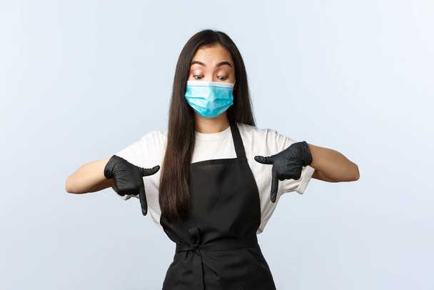 Covid-19 pandemic, social distancing, small business and preventing virus concept. Intrigued and curious asian coffee shop barista, employee in medical mask and gloves peeking down, pointing bottom - Foto, Imagen