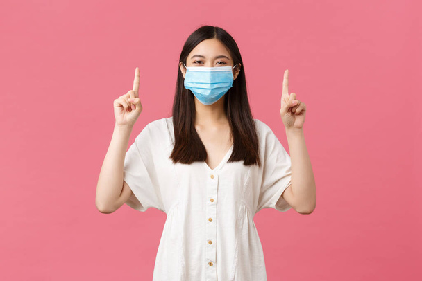 Covid-19, social distancing, virus and lifestyle concept. Cheerful smiling woman in stylish white dress, summer outfit and medical mask, pointing fingers up, promoting advertisement, pink background - Foto, Imagem