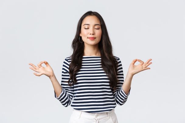 Lifestyle, people emotions and casual concept. Relaxed and patient smiling young asian woman with closed eyes meditating to calm down, do breathing exercises with hands in zen gesture - Photo, Image
