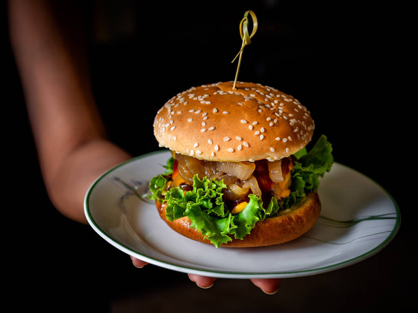 Burger maison prêt à manger. Burger juteux maison à la main avec bœuf, fromage, laitue verte fraîche et oignons caramélisés dans une assiette blanche sur fond sombre. - Photo, image