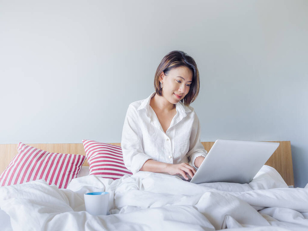 白いシャツを着て短い髪を持つ美しいアジアの女性は、コピースペースを持つ家の白い寝室で木製のベッドの上にノートパソコンで作業しています。自宅のコンセプトでリラックスして働くビジネス女性. - 写真・画像