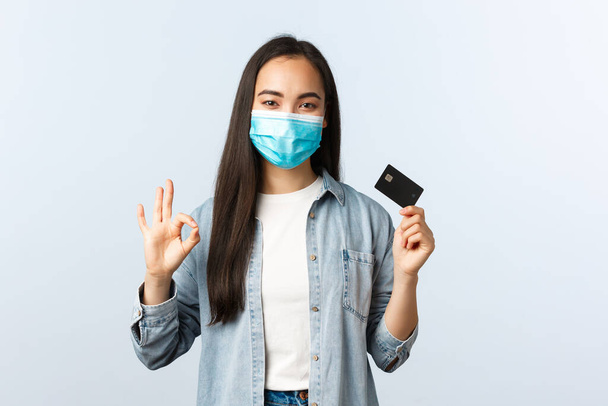 Social distancing lifestyle, covid-19 pandemic and contactless shopping concept. Satisfied asian female in medical mask show okay gesture and promote credit card features, bankc account - Foto, Imagem