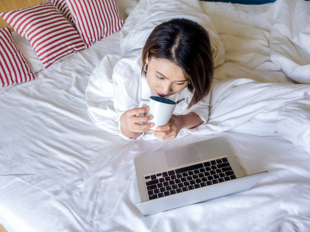 De belles femmes asiatiques aux cheveux courts portant une chemise blanche tenant une tasse de café blanche et regardant un ordinateur portable sur un lit en bois dans une chambre blanche dans la maison. - Photo, image