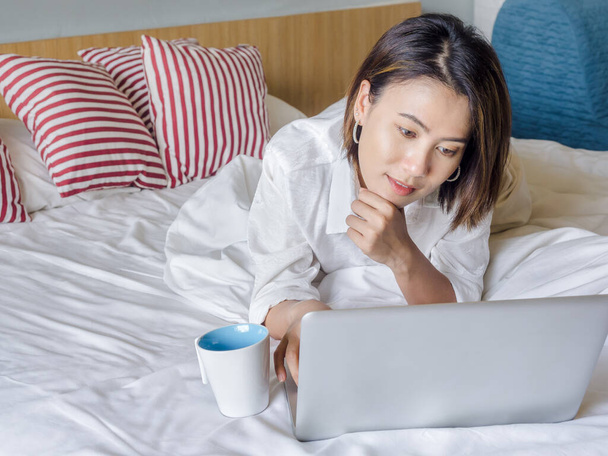 Beautiful Asian women with short hair wearing white shirt with white coffee cup and reading on laptop computer on wooden bed in white bedroom in the house. - Photo, Image