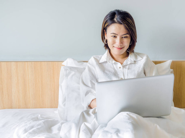 Beautiful Asian women with short hair wearing white shirt working with laptop computer on wooden bed in white bedroom in house with copy space. Business woman working with relaxation at home concept. - 写真・画像