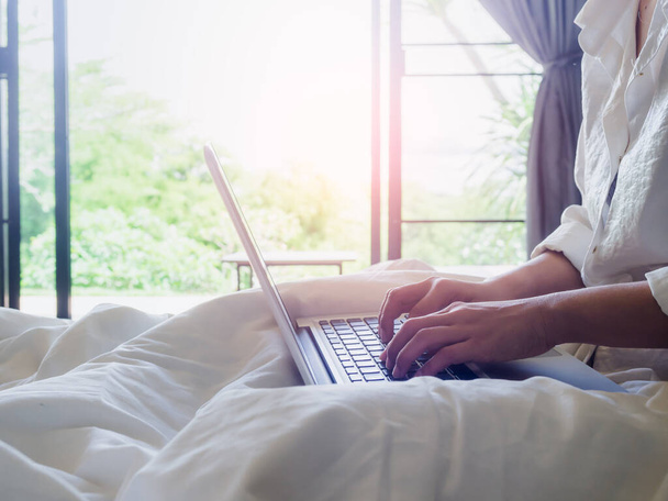 Ręce kobiety piszącej na laptopie na łóżku w domu ze światłem słonecznym z zewnątrz przez szklane drzwi. Praca w domu z relaksem. - Zdjęcie, obraz