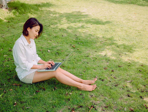 Hermosas mujeres asiáticas con camisa blanca y pantalones cortos que trabajan con ordenador portátil sentado en la hierba verde en el parque. Feliz mujer al aire libre trabajando con tecnología, educación y concepto de trabajo remoto
. - Foto, imagen
