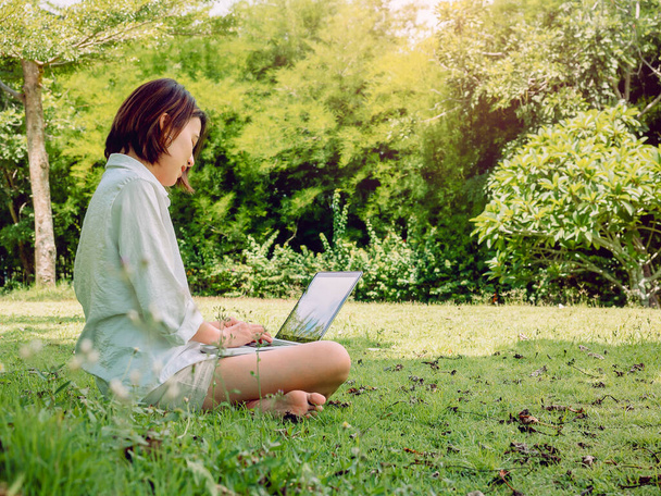 白いシャツを着た美しいアジアの女性と、公園の緑の芝生の上に座ってノートパソコンで働くショートパンツ。テクノロジー、教育、遠隔作業のコンセプトで働く幸せな女性の屋外作業. - 写真・画像