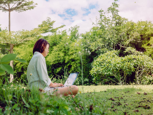 Kauniita aasialaisia naisia yllään valkoinen paita ja shortsit työskentelevät kannettavan tietokoneen istuu vihreää ruohoa puistossa. Onnellinen naisten ulkona työskentelee teknologian, koulutuksen ja etätyön konsepti. - Valokuva, kuva