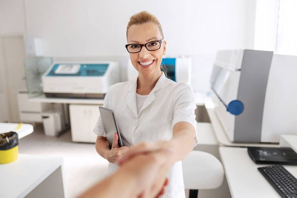 Positive friendly attractive lab assistant holding tablet under armpit and shaking hands with patient. - Foto, Imagen