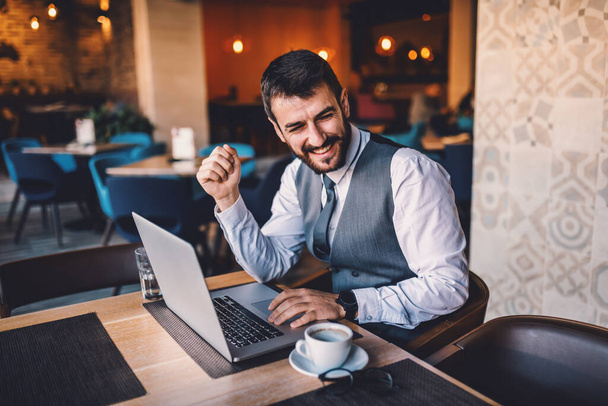 Young sophisticated caucasian satisfied bearded businessman in suit sitting in cafe, surfing on internet and looking away. On table are laptop and coffee. - Фото, изображение