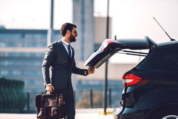 Handsome smiling positive Caucasian bearded businessman in suit with sunglasses holding his briefcase and closing trunk on his car. - Φωτογραφία, εικόνα
