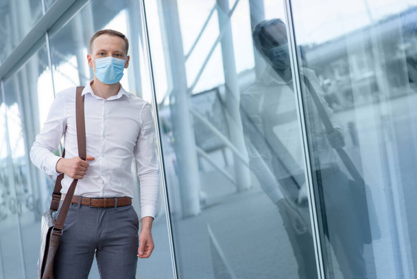 Portret van een jonge zakenman met een medisch masker. Een man staat naast de ramen van een modern kantoorgebouw. - Foto, afbeelding